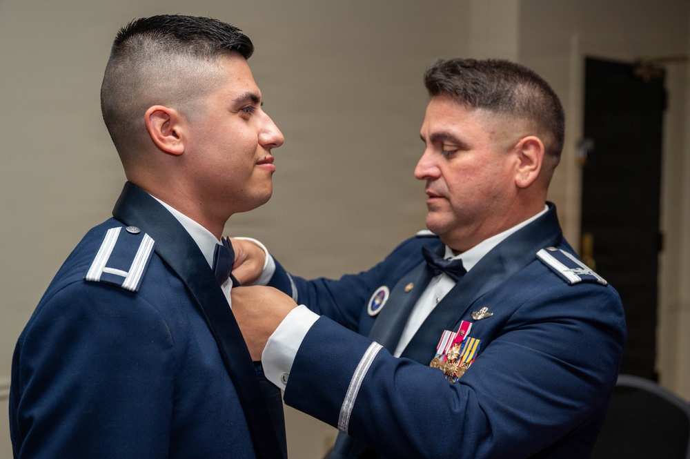 Two individuals in U.S. Air Force dress blue uniforms are engaged in what appears to be a formal military ceremony or preparation. An older, senior-ranking officer is adjusting or pinning something on the collar/chest area of a younger, junior-ranking airman. Both wear formal dress uniforms with rank insignia visible on their epaulettes.