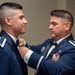 Two individuals in U.S. Air Force dress blue uniforms are engaged in what appears to be a formal military ceremony or preparation. An older, senior-ranking officer is adjusting or pinning something on the collar/chest area of a younger, junior-ranking airman. Both wear formal dress uniforms with rank insignia visible on their epaulettes.