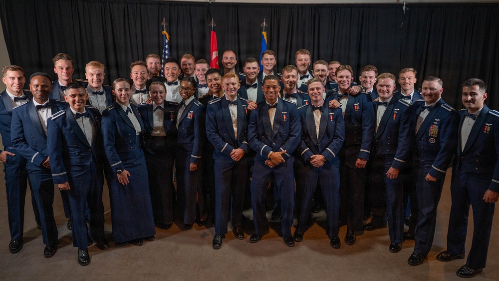 A large group of military personnel, approximately 30-35 individuals, pose together for a formal group photograph. They are dressed in dark navy blue dress uniforms and formal attire, with some members displaying medals and ribbons. Three flags are visible in the background, including the United States flag and what appears to be military branch flags.