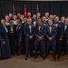 A large group of military personnel, approximately 30-35 individuals, pose together for a formal group photograph. They are dressed in dark navy blue dress uniforms and formal attire, with some members displaying medals and ribbons. Three flags are visible in the background, including the United States flag and what appears to be military branch flags.