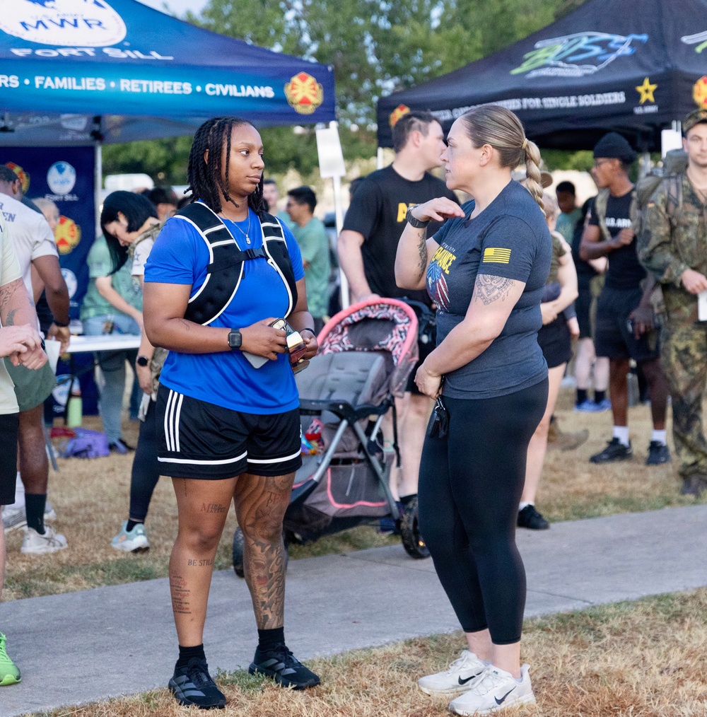 An outdoor military community event at Fort Sill, likely a fitness or wellness fair organized by MWR (Morale, Welfare, and Recreation). Two women are engaged in conversation in the foreground, while a crowd of participants including uniformed military personnel mingles in the background. Vendor tents and signage indicate this is an organized community gathering for soldiers, families, retirees, and civilians.