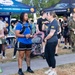 An outdoor military community event at Fort Sill, likely a fitness or wellness fair organized by MWR (Morale, Welfare, and Recreation). Two women are engaged in conversation in the foreground, while a crowd of participants including uniformed military personnel mingles in the background. Vendor tents and signage indicate this is an organized community gathering for soldiers, families, retirees, and civilians.
