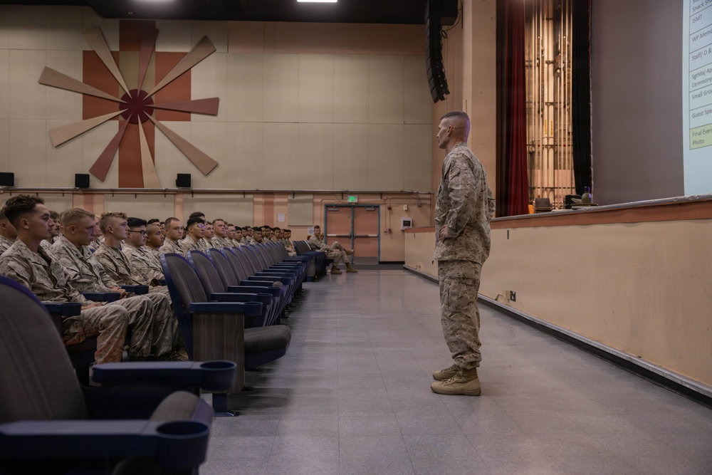 A military briefing or address is taking place in an auditorium-style venue, where a single standing service member in camouflage uniform faces a seated audience of fellow military personnel. The speaker stands at the front near a stage, addressing rows of uniformed individuals seated in blue theater-style chairs. A projected presentation is partially visible on the right side of the frame.