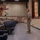 A military briefing or address is taking place in an auditorium-style venue, where a single standing service member in camouflage uniform faces a seated audience of fellow military personnel. The speaker stands at the front near a stage, addressing rows of uniformed individuals seated in blue theater-style chairs. A projected presentation is partially visible on the right side of the frame.