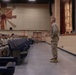 A military briefing or address is taking place in an auditorium-style venue, where a single standing service member in camouflage uniform faces a seated audience of fellow military personnel. The speaker stands at the front near a stage, addressing rows of uniformed individuals seated in blue theater-style chairs. A projected presentation is partially visible on the right side of the frame.