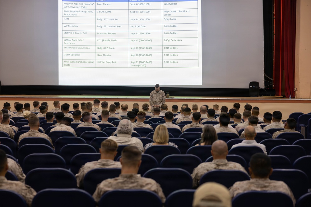 A military briefing is taking place in a theater-style auditorium, with a large projected schedule displayed on screen. A single uniformed service member stands at the front addressing rows of seated personnel, the majority wearing Marine Corps MARPAT camouflage uniforms. The projected slide appears to outline a multi-day event schedule with dates ranging from September 9–11, referencing locations and personnel including '1stLt Geddes' and 'MSgt Crow.'