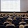 A military briefing is taking place in a theater-style auditorium, with a large projected schedule displayed on screen. A single uniformed service member stands at the front addressing rows of seated personnel, the majority wearing Marine Corps MARPAT camouflage uniforms. The projected slide appears to outline a multi-day event schedule with dates ranging from September 9–11, referencing locations and personnel including '1stLt Geddes' and 'MSgt Crow.'