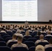 A military briefing is taking place in a theater-style auditorium, with a large projected schedule displayed on screen. A single uniformed service member stands at the front addressing rows of seated personnel, the majority wearing Marine Corps MARPAT camouflage uniforms. The projected slide appears to outline a multi-day event schedule with dates ranging from September 9–11, referencing locations and personnel including '1stLt Geddes' and 'MSgt Crow.'