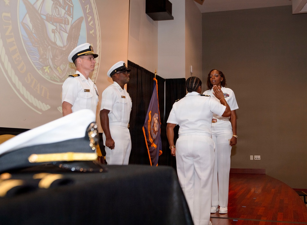 A formal United States Navy ceremony is taking place in an indoor venue. Four personnel in white Navy dress uniforms are visible, with a projected U.S. Navy seal on a screen in the background. The scene appears to depict a change of command or similar official military ceremony.