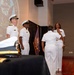 A formal United States Navy ceremony is taking place in an indoor venue. Four personnel in white Navy dress uniforms are visible, with a projected U.S. Navy seal on a screen in the background. The scene appears to depict a change of command or similar official military ceremony.