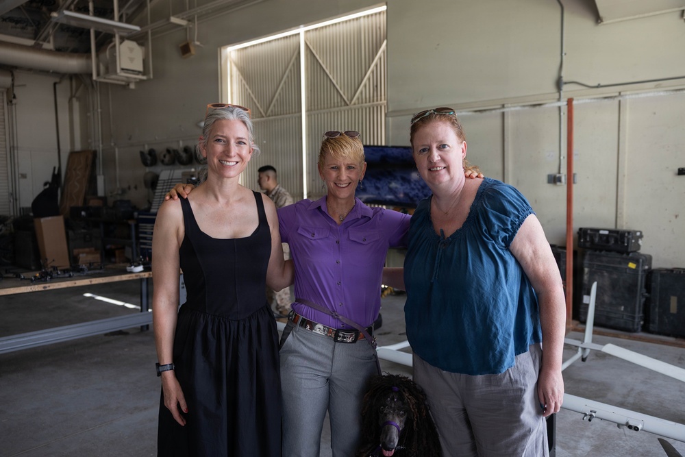 Three women are posing together for a photo inside what appears to be a large industrial or military hangar facility. A dark-colored dog, possibly a Standard Poodle, is visible between them in the foreground. The setting contains various equipment, cases, and workbenches in the background.