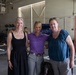 Three women are posing together for a photo inside what appears to be a large industrial or military hangar facility. A dark-colored dog, possibly a Standard Poodle, is visible between them in the foreground. The setting contains various equipment, cases, and workbenches in the background.