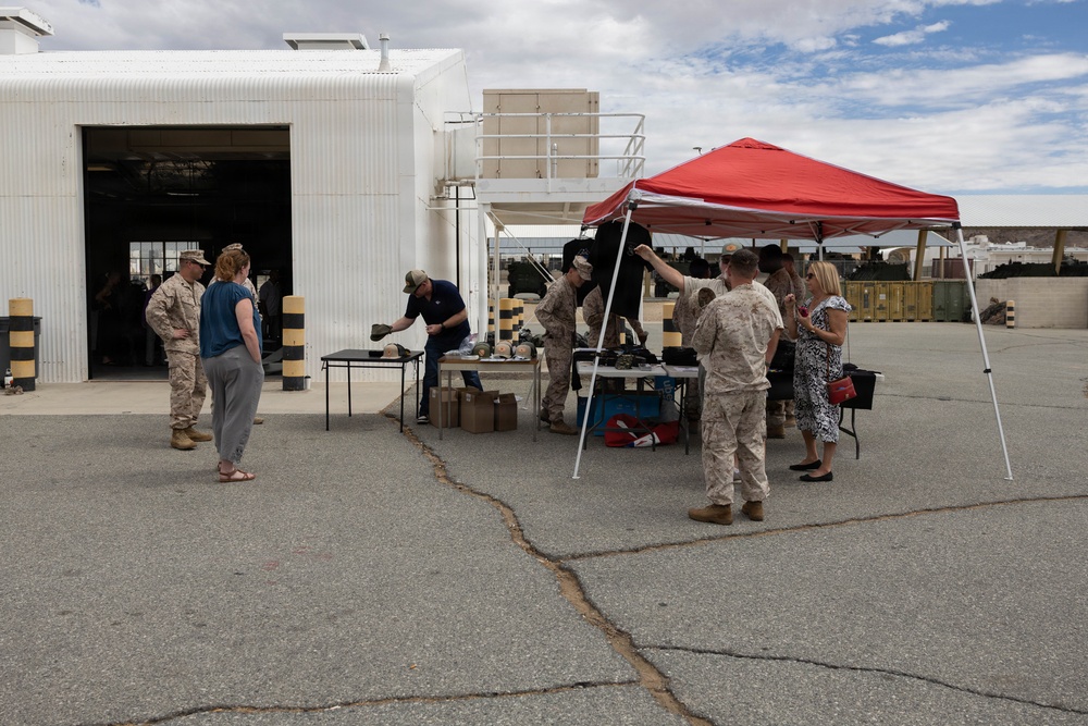 A group of military personnel in camouflage uniforms and civilians gather around tables set up under a red pop-up canopy tent in an outdoor paved area near a white industrial building. The setting appears to be a military installation or base, with items displayed on the tables suggesting a demonstration, equipment display, or informational event. Both uniformed Marines and civilian attendees, including women in casual clothing, are actively engaged at the tables.