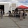 A group of military personnel in camouflage uniforms and civilians gather around tables set up under a red pop-up canopy tent in an outdoor paved area near a white industrial building. The setting appears to be a military installation or base, with items displayed on the tables suggesting a demonstration, equipment display, or informational event. Both uniformed Marines and civilian attendees, including women in casual clothing, are actively engaged at the tables.