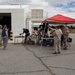 A group of military personnel in camouflage uniforms and civilians gather around tables set up under a red pop-up canopy tent in an outdoor paved area near a white industrial building. The setting appears to be a military installation or base, with items displayed on the tables suggesting a demonstration, equipment display, or informational event. Both uniformed Marines and civilian attendees, including women in casual clothing, are actively engaged at the tables.