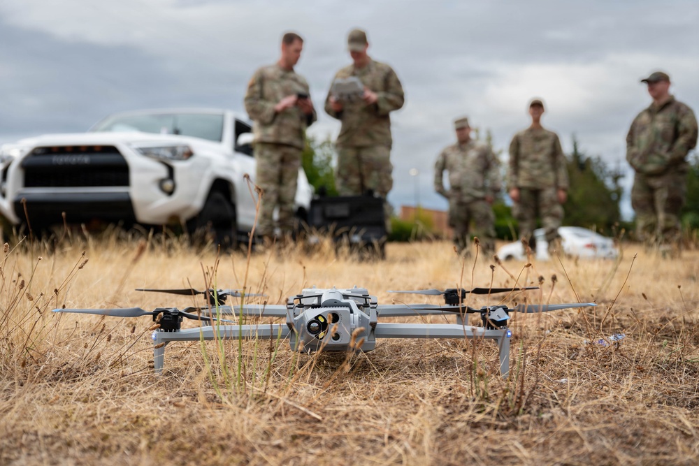 A military quadcopter drone sits on dry grass in the foreground while five personnel in camouflage uniforms stand in the background. Two of the personnel appear to be reviewing documents or operating a tablet/controller, while others observe. A white Toyota 4Runner SUV and equipment cases are visible behind the group.