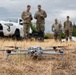 A military quadcopter drone sits on dry grass in the foreground while five personnel in camouflage uniforms stand in the background. Two of the personnel appear to be reviewing documents or operating a tablet/controller, while others observe. A white Toyota 4Runner SUV and equipment cases are visible behind the group.