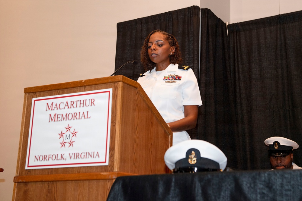 A female U.S. Navy officer in white service dress uniform is speaking at a podium during an official ceremony at the MacArthur Memorial in Norfolk, Virginia. She displays multiple service ribbons on her uniform, indicating significant military experience. In the foreground and background, additional Navy personnel in white uniforms are visible.