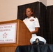 A female U.S. Navy officer in white service dress uniform is speaking at a podium during an official ceremony at the MacArthur Memorial in Norfolk, Virginia. She displays multiple service ribbons on her uniform, indicating significant military experience. In the foreground and background, additional Navy personnel in white uniforms are visible.