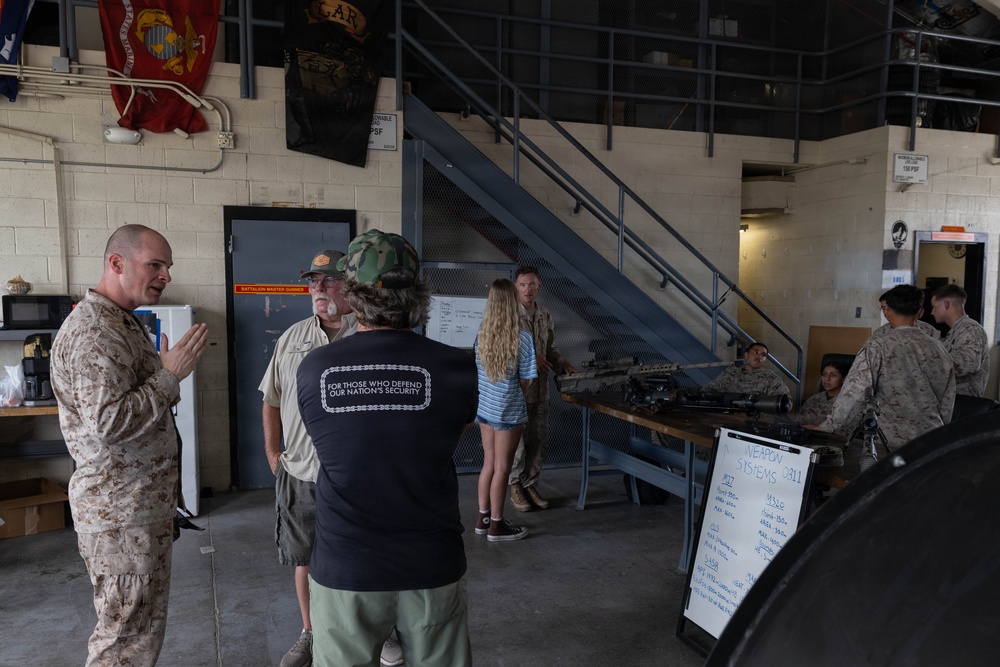 A group of civilians is being given a tour or briefing at what appears to be a U.S. Marine Corps facility. A Marine in desert camouflage uniform is speaking to visitors, while in the background military weapons are displayed on a table with a whiteboard labeled 'Weapon Systems 0311'. Military flags and unit insignia are visible on the walls, suggesting this is an official visit or community outreach event.