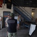 A group of civilians is being given a tour or briefing at what appears to be a U.S. Marine Corps facility. A Marine in desert camouflage uniform is speaking to visitors, while in the background military weapons are displayed on a table with a whiteboard labeled 'Weapon Systems 0311'. Military flags and unit insignia are visible on the walls, suggesting this is an official visit or community outreach event.