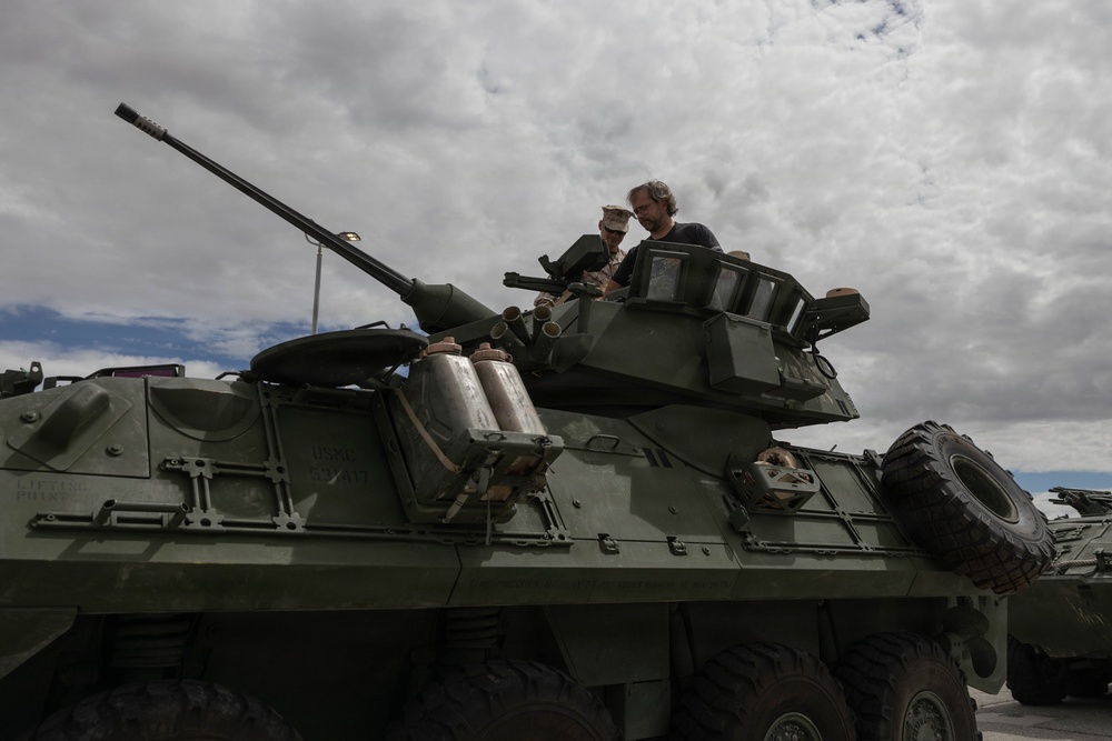Two individuals are positioned in the turret hatch of a military armored vehicle, appearing to examine or discuss equipment. The vehicle is an olive-drab wheeled armored fighting vehicle, consistent with a Light Armored Vehicle (LAV) type, equipped with a cannon and smoke grenade launchers. The scene appears to be a static display or inspection setting under an overcast sky.