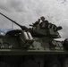 Two individuals are positioned in the turret hatch of a military armored vehicle, appearing to examine or discuss equipment. The vehicle is an olive-drab wheeled armored fighting vehicle, consistent with a Light Armored Vehicle (LAV) type, equipped with a cannon and smoke grenade launchers. The scene appears to be a static display or inspection setting under an overcast sky.