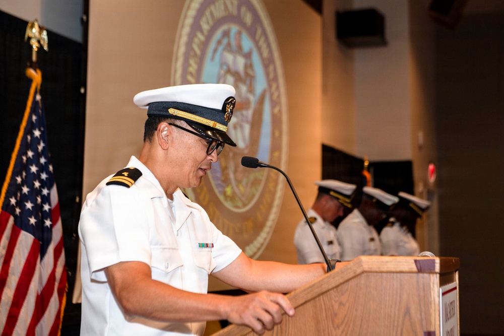 A U.S. Navy officer in white service dress uniform is speaking at a podium during what appears to be an official military ceremony. The Department of the Navy seal is projected on the wall behind the speaker, and additional uniformed personnel are visible in the background. An American flag is displayed to the left of the podium.