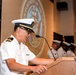 A U.S. Navy officer in white service dress uniform is speaking at a podium during what appears to be an official military ceremony. The Department of the Navy seal is projected on the wall behind the speaker, and additional uniformed personnel are visible in the background. An American flag is displayed to the left of the podium.