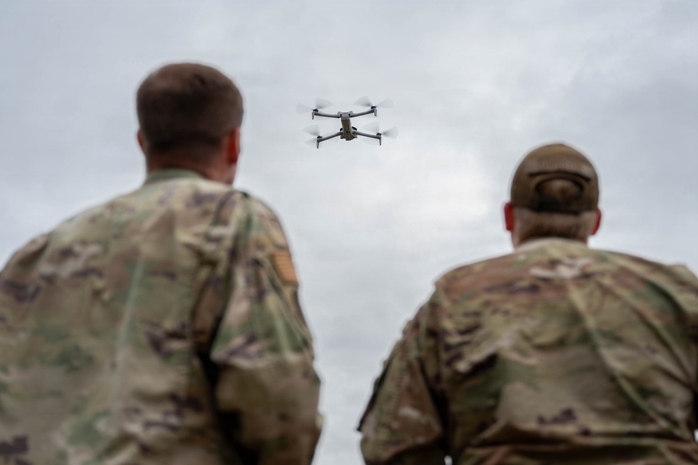 Two military personnel in camouflage uniforms are observed from behind, watching a small quadcopter drone hovering in an overcast sky. The drone appears to be a commercial-style quadcopter, possibly used for reconnaissance or training purposes. The image captures what appears to be a drone operation or training exercise.