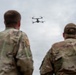 Two military personnel in camouflage uniforms are observed from behind, watching a small quadcopter drone hovering in an overcast sky. The drone appears to be a commercial-style quadcopter, possibly used for reconnaissance or training purposes. The image captures what appears to be a drone operation or training exercise.