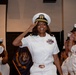 A U.S. Navy officer in white dress uniform is saluting during what appears to be a formal military ceremony. She is smiling broadly and wearing a white officer's cover with gold insignia, multiple service ribbons, and rank insignia on her collar. Several other Navy personnel in white uniforms are visible in the background, some also saluting.