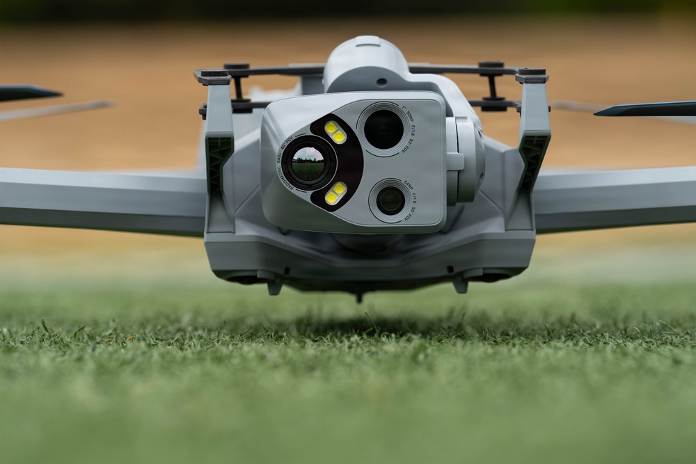 A close-up front-facing photograph of a consumer or professional-grade quadcopter drone resting on a grass surface. The drone features a multi-sensor camera gimbal system with multiple lenses and LED lights visible on the front payload. The image is taken at ground level, providing a dramatic low-angle perspective of the aircraft.