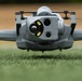 A close-up front-facing photograph of a consumer or professional-grade quadcopter drone resting on a grass surface. The drone features a multi-sensor camera gimbal system with multiple lenses and LED lights visible on the front payload. The image is taken at ground level, providing a dramatic low-angle perspective of the aircraft.