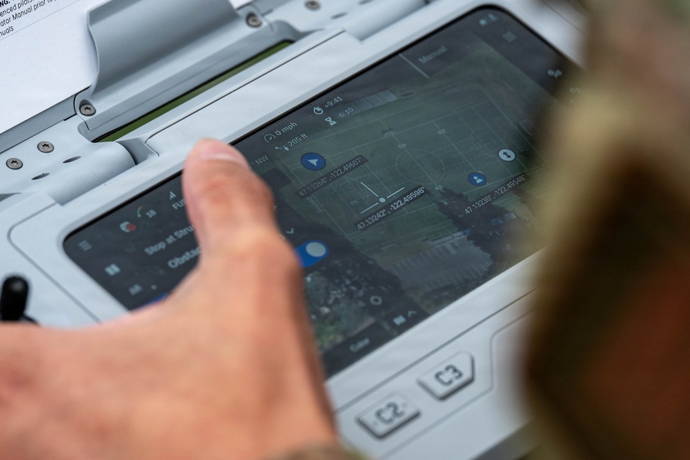 A person is interacting with a ruggedized tablet or military-grade computing device displaying a drone or UAV ground control station interface. The screen shows a satellite map view with GPS coordinates, flight path waypoints, and telemetry data. The device appears to be a Panasonic Toughbook or similar rugged field computer used for unmanned aerial vehicle operations.