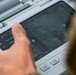 A person is interacting with a ruggedized tablet or military-grade computing device displaying a drone or UAV ground control station interface. The screen shows a satellite map view with GPS coordinates, flight path waypoints, and telemetry data. The device appears to be a Panasonic Toughbook or similar rugged field computer used for unmanned aerial vehicle operations.