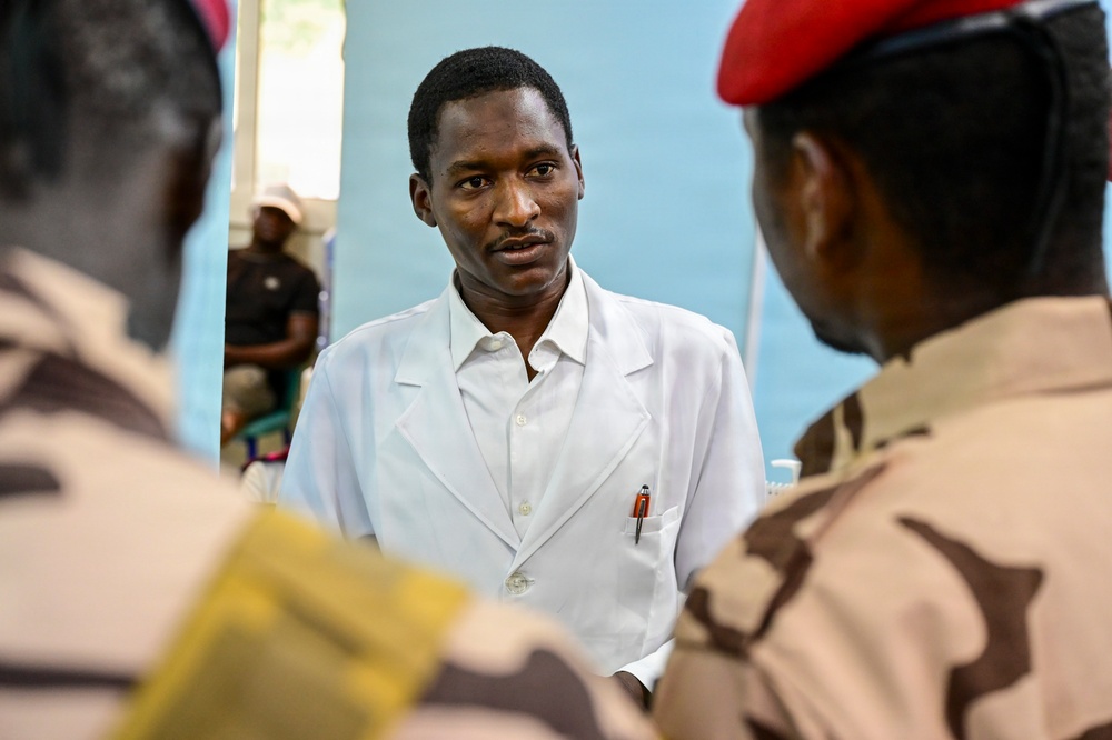 A man wearing a white medical coat with a pen in the breast pocket appears to be speaking with two individuals in desert camouflage military uniforms, one of whom is wearing a red beret. The setting appears to be an indoor medical or administrative facility with a light blue wall visible in the background. A fourth individual in civilian clothing is seated in the background.