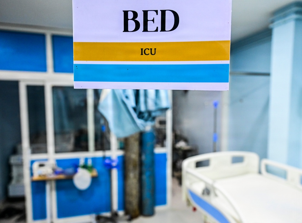 The image shows an ICU (Intensive Care Unit) bed area in a hospital setting. A sign in the foreground clearly reads 'BED ICU' with yellow and blue color-coded stripes. The background reveals a partially visible hospital bed, medical equipment, and blue-accented room partitions.