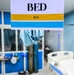The image shows an ICU (Intensive Care Unit) bed area in a hospital setting. A sign in the foreground clearly reads 'BED ICU' with yellow and blue color-coded stripes. The background reveals a partially visible hospital bed, medical equipment, and blue-accented room partitions.