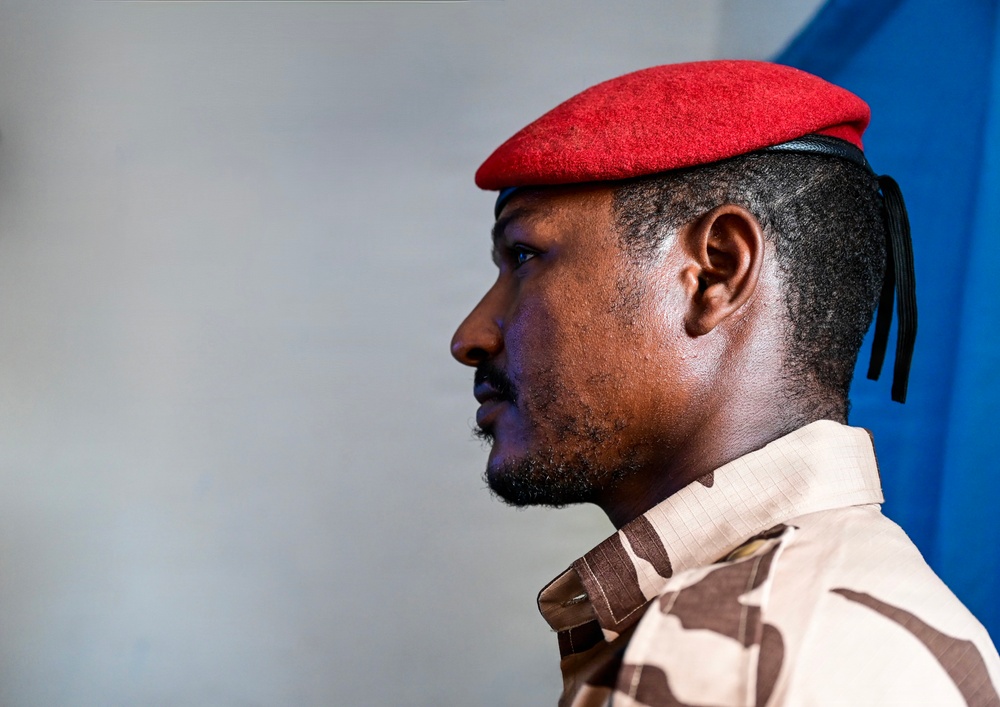 A profile portrait of a male soldier wearing a red beret and desert camouflage uniform. The image is a close-up side view showing strong facial features and military attire. The background is a neutral gray with a partial blue element visible on the right edge.