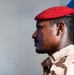 A profile portrait of a male soldier wearing a red beret and desert camouflage uniform. The image is a close-up side view showing strong facial features and military attire. The background is a neutral gray with a partial blue element visible on the right edge.