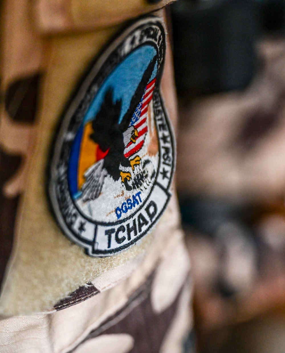 The image shows a military or security force patch attached via velcro to a desert camouflage uniform or vest. The patch references 'TCHAD' (French for Chad) and includes the acronym 'DGSAT', along with an eagle motif and what appears to be an American flag, suggesting a joint or advisory mission in Chad. The patch is circular with a banner at the bottom and stars around the border.
