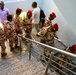 A group of uniformed military or paramilitary personnel wearing desert camouflage uniforms and distinctive red berets are gathered on a staircase landing inside a modern building. They appear to be moving as a group, with some ascending and others congregating near a stainless steel railing. Two civilians in casual clothing are also visible in the background.