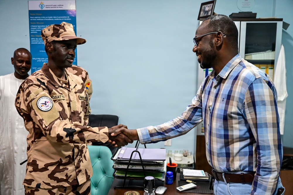 A military officer in desert camouflage uniform is shaking hands with a civilian man wearing a plaid shirt in what appears to be an office setting. A third person in white traditional clothing is visible in the background. The setting appears to be an official meeting or formal greeting, likely in an African country based on contextual clues.