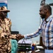 A military officer in desert camouflage uniform is shaking hands with a civilian man wearing a plaid shirt in what appears to be an office setting. A third person in white traditional clothing is visible in the background. The setting appears to be an official meeting or formal greeting, likely in an African country based on contextual clues.