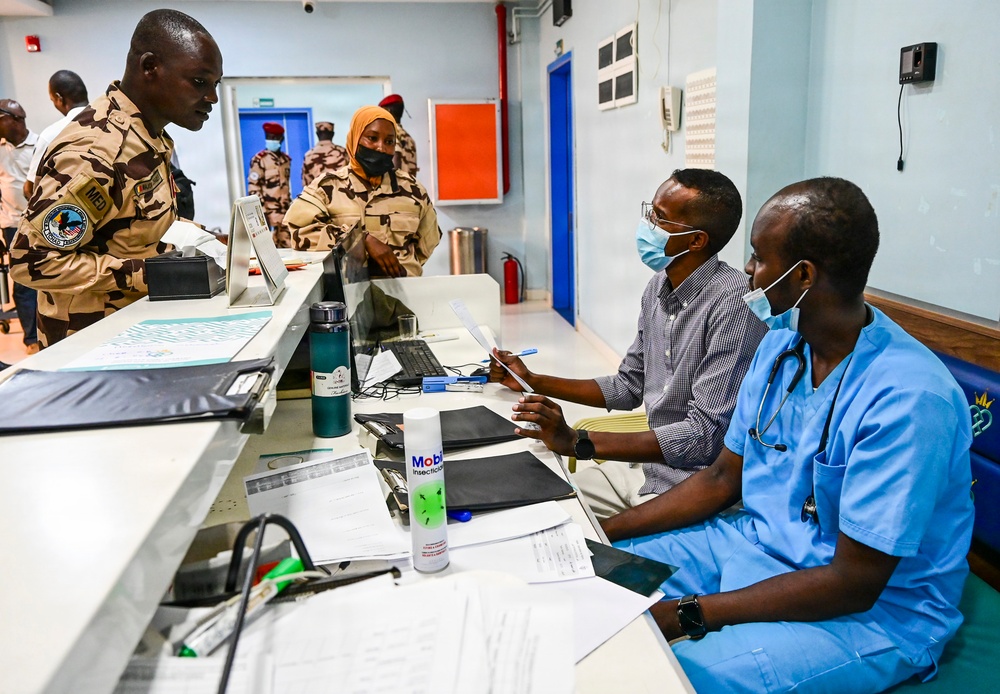 The image shows a medical facility where military personnel in desert camouflage uniforms are interacting with healthcare workers at a reception or triage desk. Two medical staff members, one wearing blue scrubs with a stethoscope and both wearing face masks, appear to be consulting with or processing the uniformed individuals. The setting appears to be a clinic or hospital intake area with administrative materials and medical supplies visible on the desk.