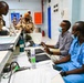 The image shows a medical facility where military personnel in desert camouflage uniforms are interacting with healthcare workers at a reception or triage desk. Two medical staff members, one wearing blue scrubs with a stethoscope and both wearing face masks, appear to be consulting with or processing the uniformed individuals. The setting appears to be a clinic or hospital intake area with administrative materials and medical supplies visible on the desk.