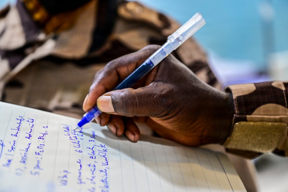 A close-up photograph of a person's hand writing in a lined notebook using a blue ballpoint pen. The individual is wearing a camouflage uniform, suggesting a military or paramilitary context. The notebook contains handwritten text in blue ink, partially legible.