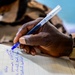 A close-up photograph of a person's hand writing in a lined notebook using a blue ballpoint pen. The individual is wearing a camouflage uniform, suggesting a military or paramilitary context. The notebook contains handwritten text in blue ink, partially legible.