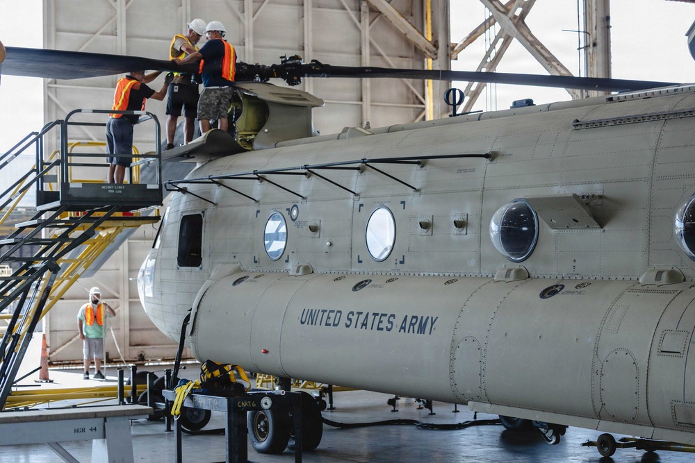 Maintenance personnel are working on a United States Army CH-47 Chinook helicopter inside a large hangar facility. Three workers wearing orange safety vests and hard hats are positioned on an elevated work platform accessing the rotor head assembly at the top of the aircraft. A fourth worker in an orange vest is visible on the hangar floor in the background.